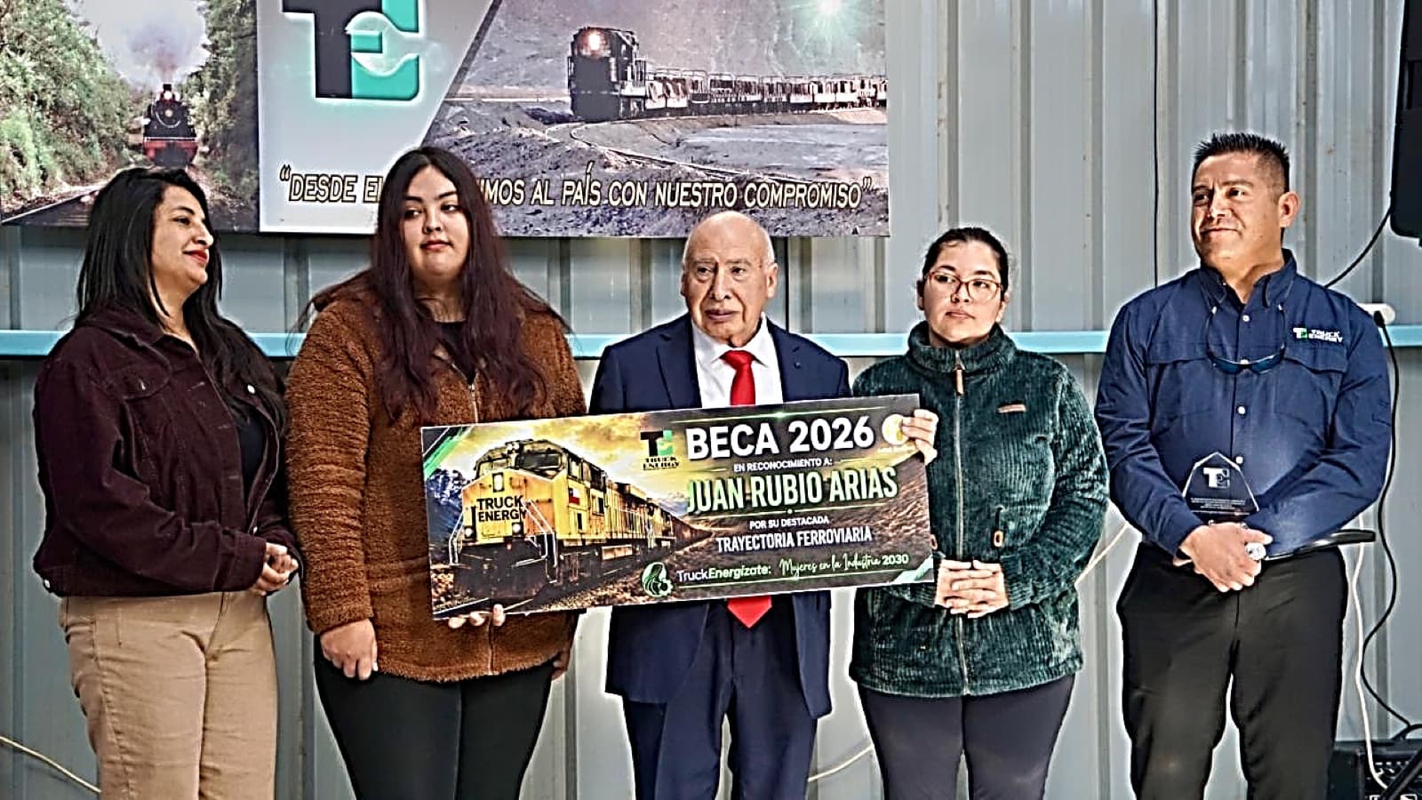 Entrega de la Beca 2026 en reconocimiento a Juan Rubio Arias: cinco personas sostienen un cheque simbólico con el nombre de la beca frente a un pendón de Truck Energy