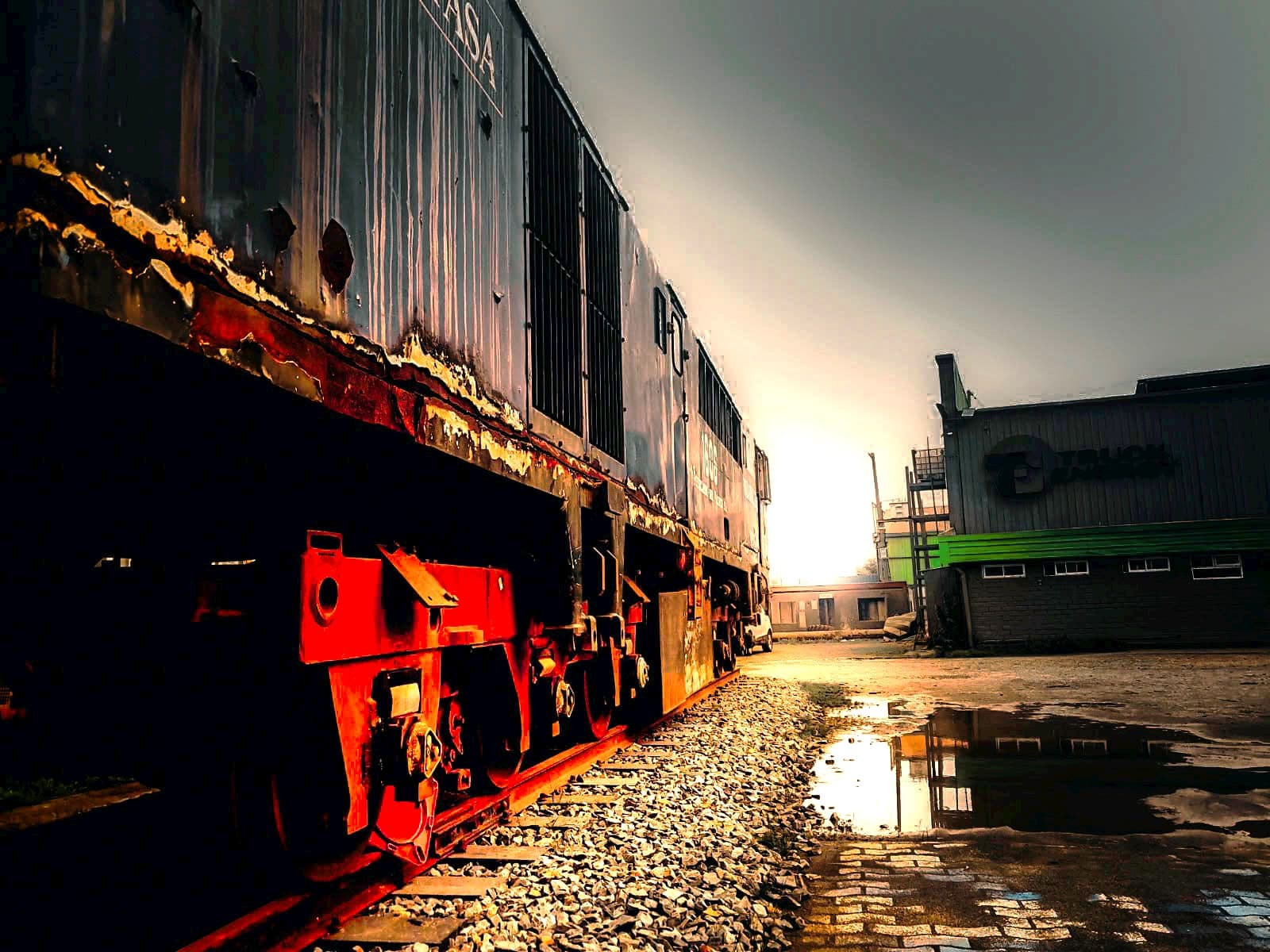 Dos locomotoras históricas detenidas sobre la vía en el patio de Truck Energy al atardecer, con la carrocería oxidada y el galpón de la empresa a la derecha