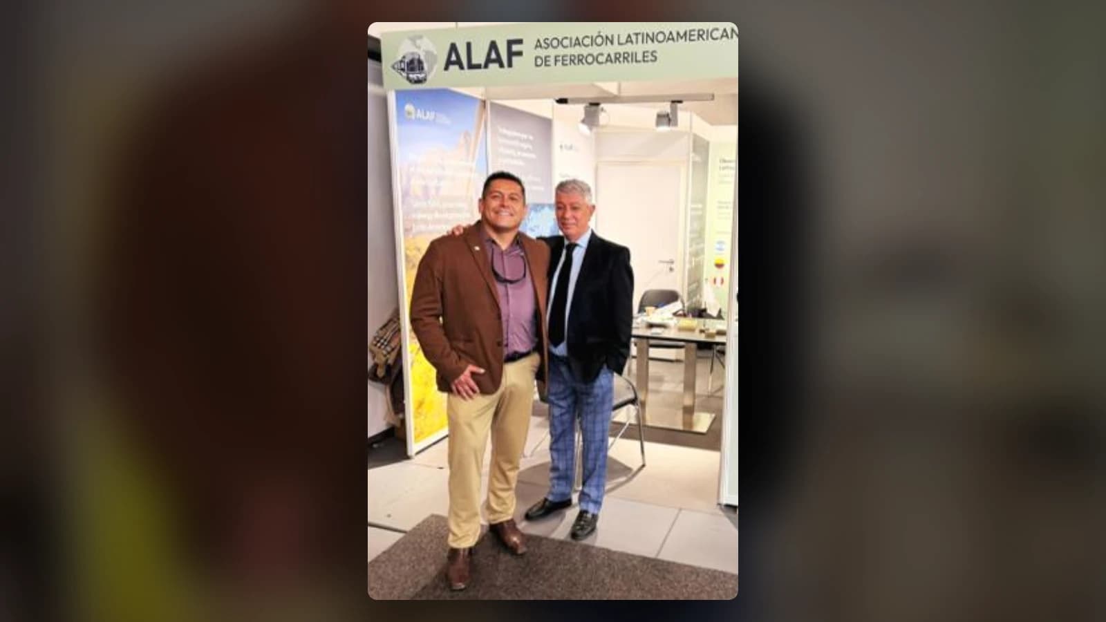 Dos hombres posan frente al stand de la Asociación Latinoamericana de Ferrocarriles (ALAF): uno con chaqueta café y pantalón beige, el otro con chaqueta oscura, corbata y pantalón a cuadros