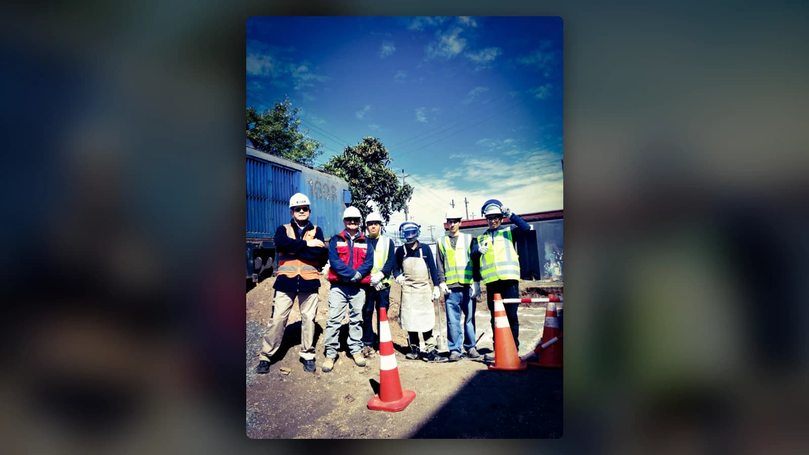 Six people in helmets and protective gear posing next to an excavation, between safety cones: three in yellow INACAP high-visibility vests, one with a face shield and a leather apron, one in a red INACAP vest and another in an orange vest. Behind them, a blue locomotive numbered 1608