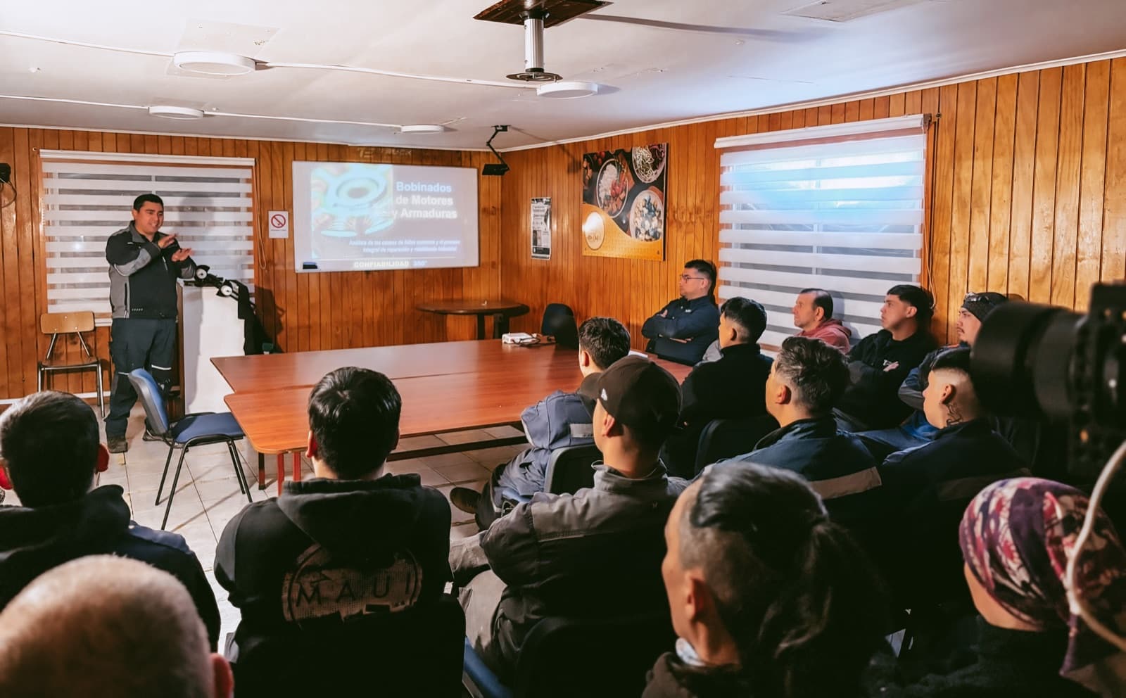 Sala de reuniones del taller con los trabajadores sentados de espaldas y el expositor de pie junto a la pantalla, donde se proyecta la lámina titulada Bobinados de Motores y Armaduras