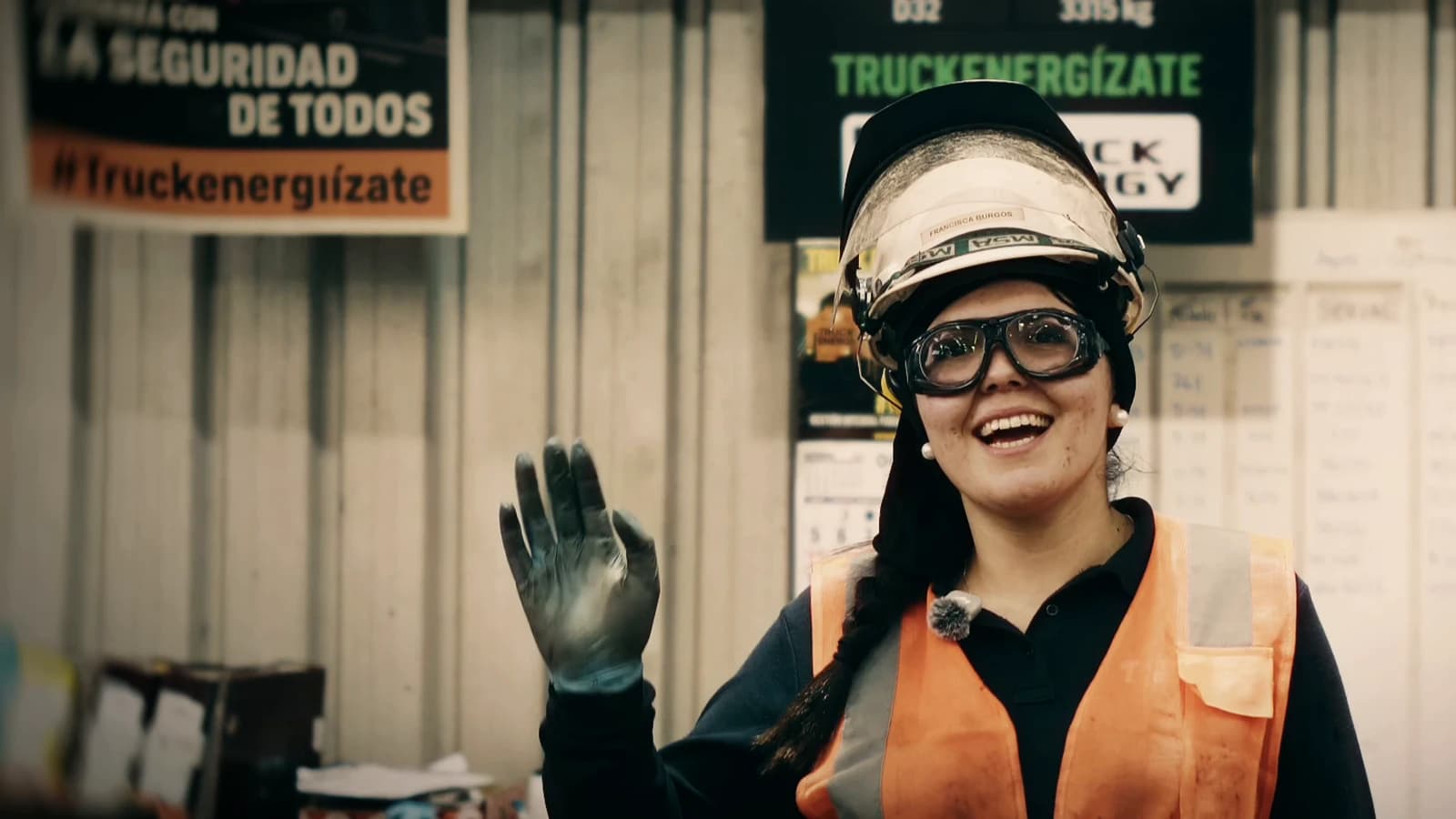 Francisca Burgos saluda con la mano y sonríe a la cámara en el taller, con casco, antiparras y chaleco reflectante, bajo el letrero Truckenergízate y junto a un cartel de seguridad con el lema #Truckenergízate