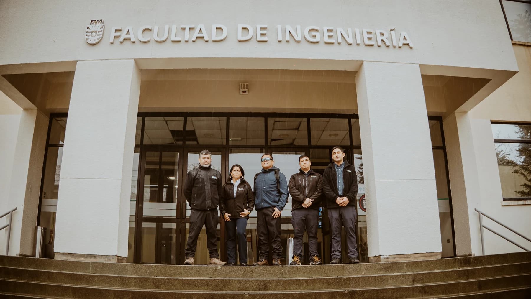 Cinco integrantes de Truck Energy de pie en la escalinata de acceso, bajo el letrero Facultad de Ingeniería y el escudo de la Universidad del Bío-Bío